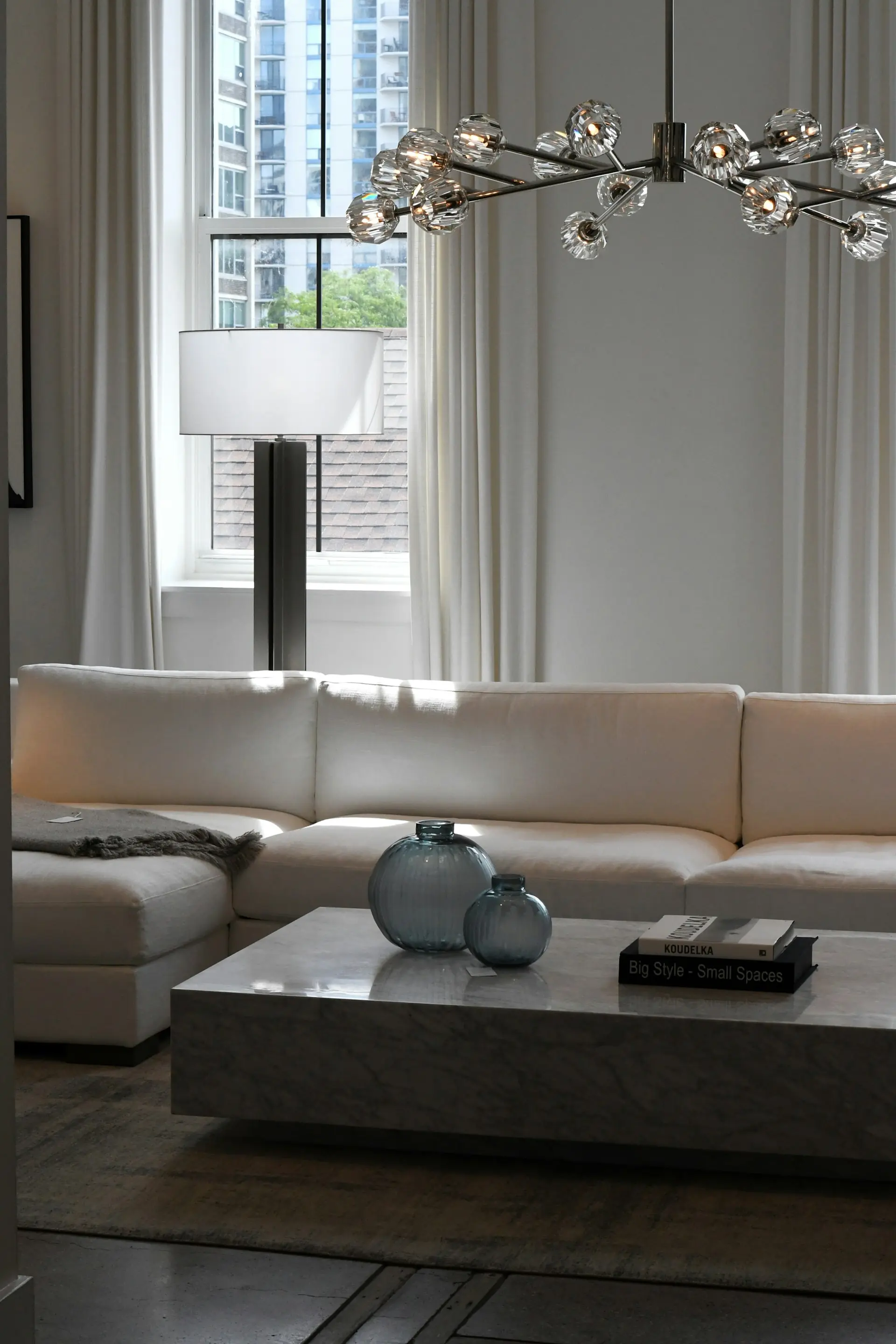 A showroom floor vignette in daylight — a white sectional and marble table under a polished-nickel chandelier, sample tags still on the pieces